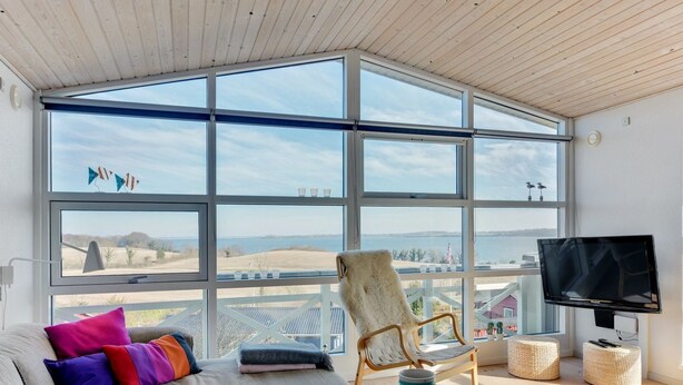 Ein geräumiges Wohnzimmer mit einem großen Fenster, das Meer und Hügel zeigt, eine bequeme Couch mit bunten Kissen, ein Holzstuhl, ein kleiner Tisch mit einer Vase und Gläsern sowie ein Fernseher an der Wand.