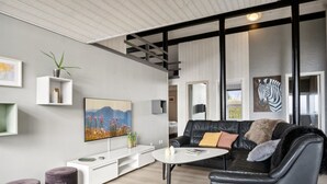 A modern living room with a black leather sofa, a white coffee table, and a mounted TV. There are two small purple ottomans and a white console table with a plant and decorative items.