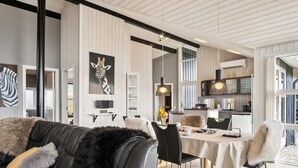 A modern living room with a black leather sofa, a dining table with chairs, and a kitchen area with a large window.