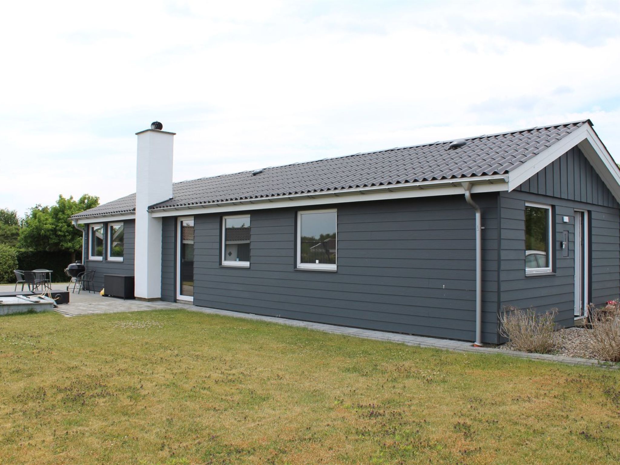 A modern single-story house with a grey exterior, a chimney, and a patio area with outdoor furniture.