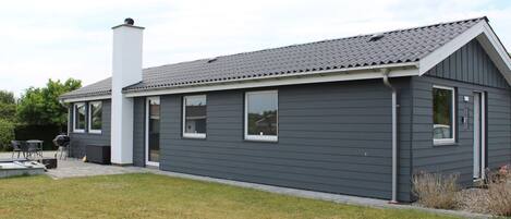 A modern single-story house with a grey exterior, a chimney, and a patio area with outdoor furniture.