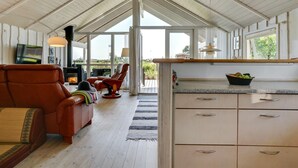 Ein großzügiger Wohnbereich mit hoher Decke, Holzbalken und großen Fenstern. Es gibt ein Ledersofa, einen Esstisch mit Obstschale sowie eine Küchenzeile mit Schränken.