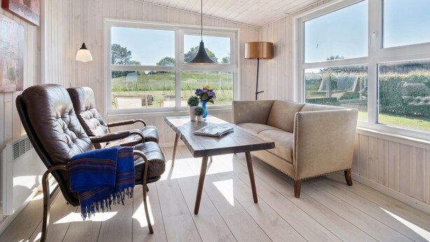 A cozy room with a wooden ceiling, large windows, a leather chair, a sofa, and a table with a vase.