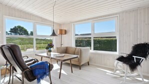 A room with a wooden ceiling, large windows, a table, a sofa, a rocking chair, and a lamp.