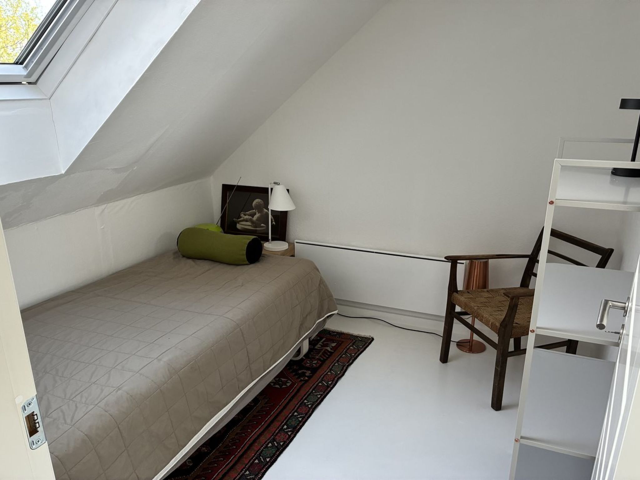 A small, well-lit room with a sloped ceiling, a single bed, a chair, a desk, and a bookshelf.