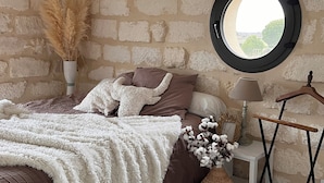 A bedroom with a round mirror, a bed with brown and white bedding, a bedside table with a lamp, and a wall-mounted shelf with decorative items.