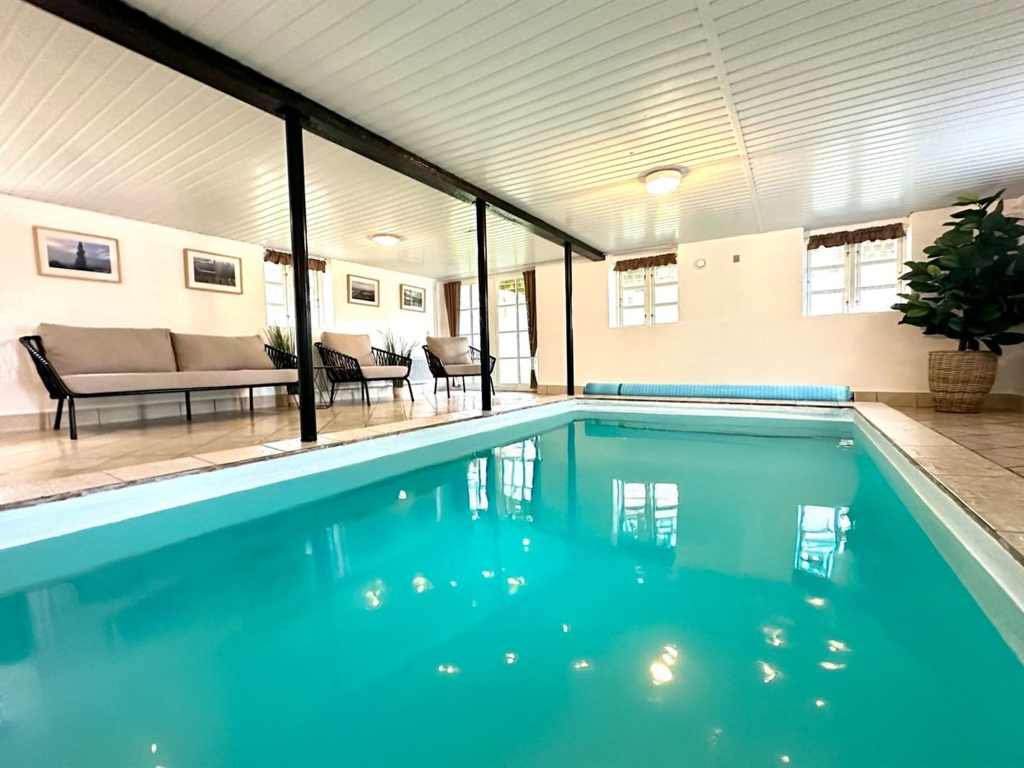Une piscine intérieure avec un sol carrelé, entourée d’un canapé, de fauteuils et d’une plante en pot.