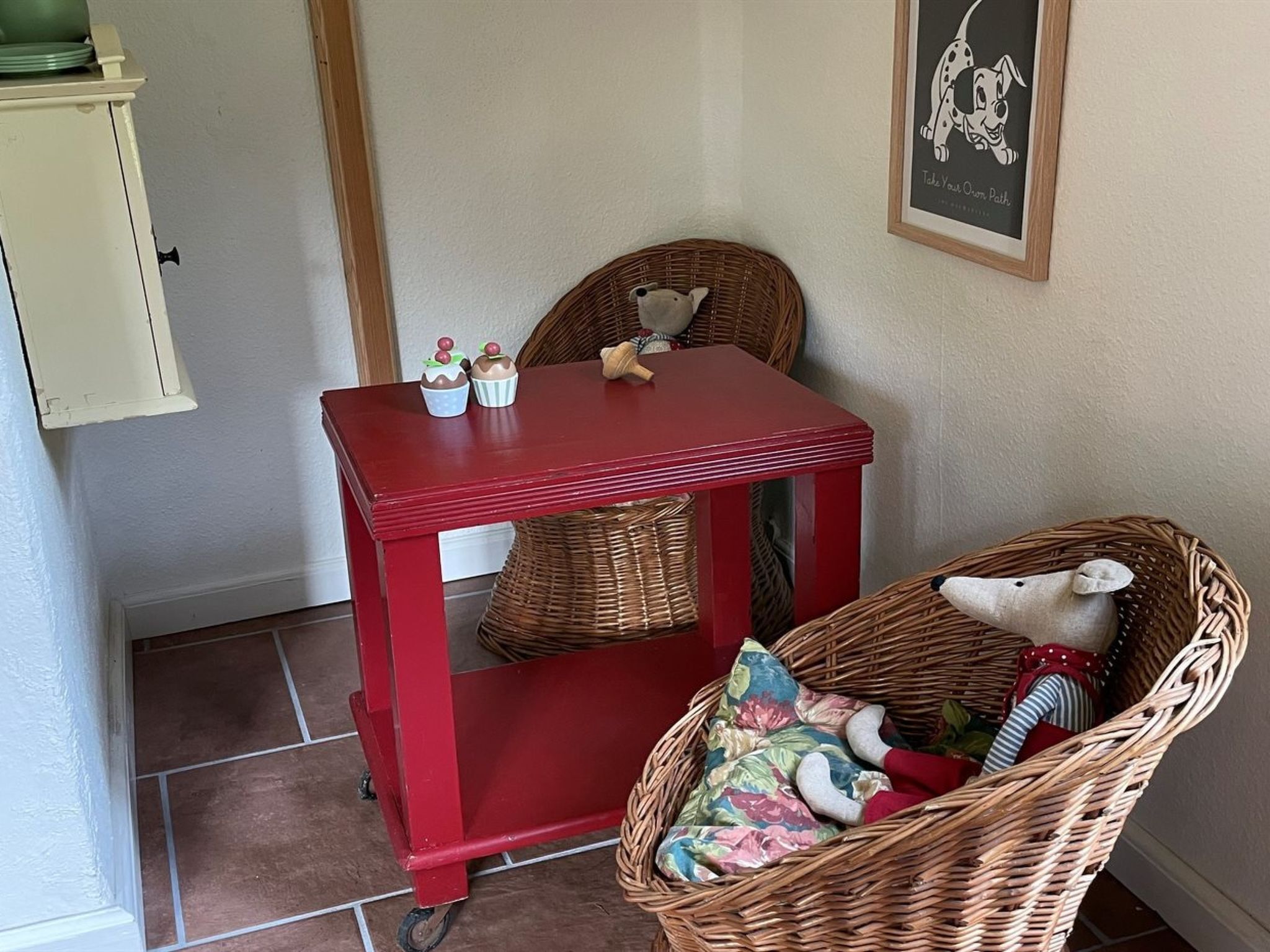 Una mesa roja con una silla de mimbre a su lado, una cesta de mimbre con un peluche, y un cuadro colgado en la pared.