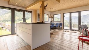 Ett rymligt kök med en central köksö, stora fönster med utsikt över bergen och en matplats med en röd stol.