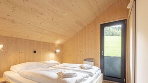 Ett sovrum i en stuga med träpanel, en säng, två vägglampor och en dörr som leder ut till en balkong med utsikt över en gräsyta.