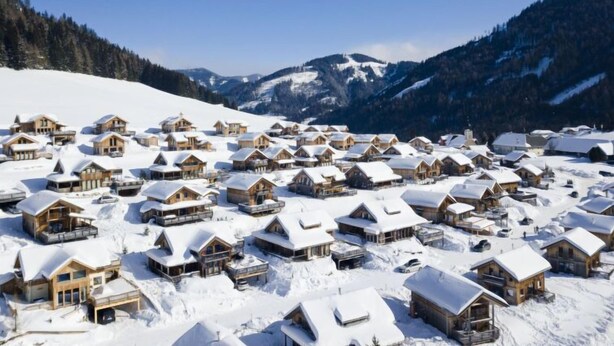 A snow-covered village with wooden chalets, a mountain backdrop, and clear skies.
