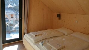 A room with a wooden sloped ceiling, a bed with two mattresses, a window with a view of buildings, and a curtain.