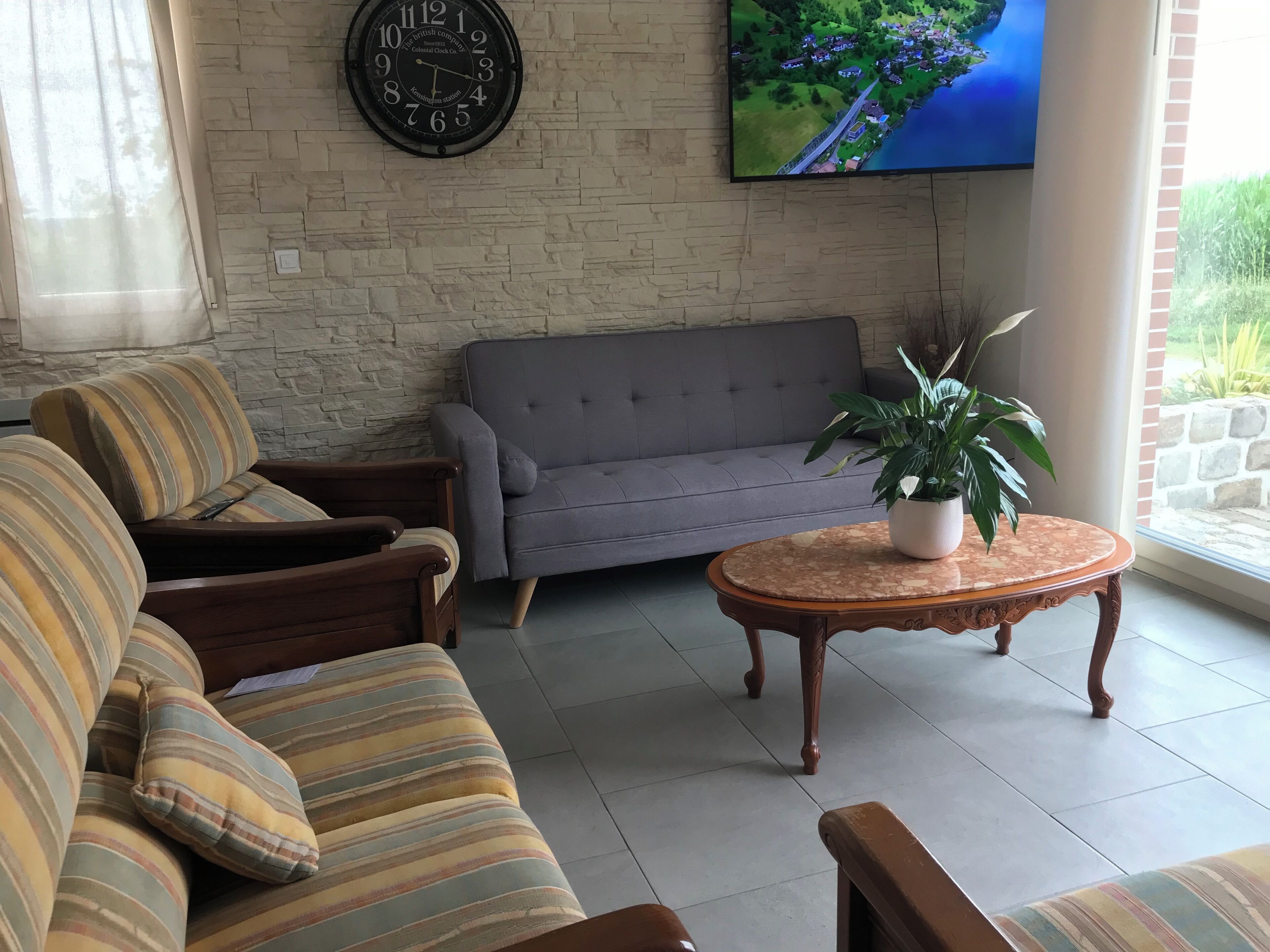 Un salon avec un canapé gris, des fauteuils à rayures, une table basse en bois sur laquelle est posée une plante en pot, une horloge murale et un téléviseur qui affiche une vue aérienne.