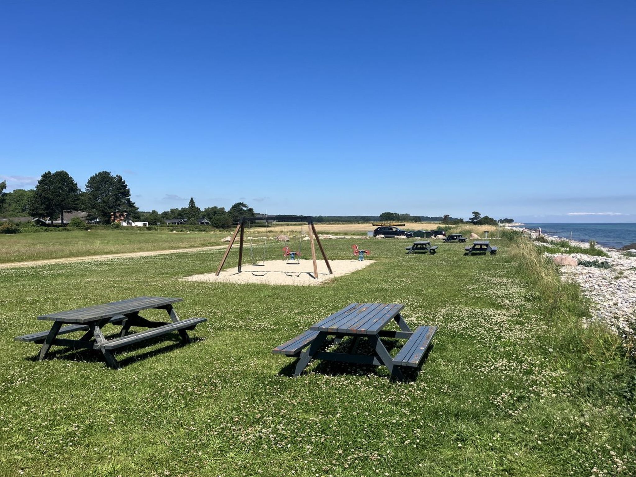 A picnic area with benches and swings, overlooking a beach with cars and a clear sky.