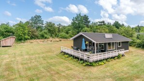 Une petite cabane à un seul étage, dotée d’un porche, entourée d’une cour verdoyante et d’arbres denses.
