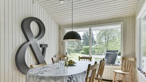 Un espace repas avec une grande table, des chaises en bois, un luminaire suspendu noir et une vue sur l’extérieur.