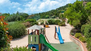 Un parque infantil con toboganes y estructuras para escalar, rodeado de follaje y colinas.
