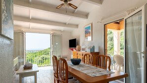 Un comedor con una mesa de madera, sillas y un banco. Hay un televisor sobre un mueble bajo, una ventana con vistas a zonas verdes y una puerta que da a un balcón.