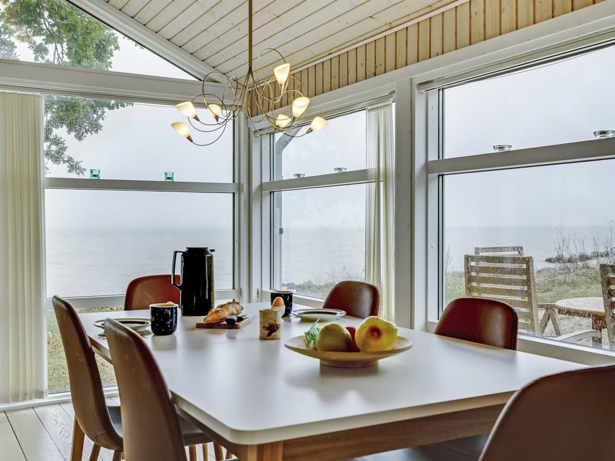 Un coin repas avec une table dressée pour deux personnes, une cafetière et un panier de fruits. De grandes fenêtres offrent une vue sur la mer.