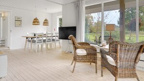 Un amplio espacio de estar con comedor, sillas de mimbre, televisor de pantalla plana y grandes ventanales que ofrecen vistas al exterior.