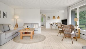 Amplia sala de estar con sofá, mesa de centro y sillas de mimbre. Hay un comedor con mesa y sillas, y un televisor montado en la pared.