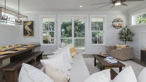 A spacious living room with a large window, a ceiling fan, and a dining area with a long table and benches. - Luxury Hilton Head Marsh View Home | Pool & Kids' Loft (Hilton Head Island)