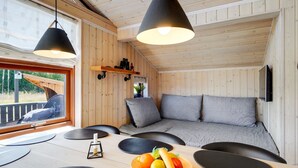 Un acogedor interior de cabaña con paredes de madera, un sofá con cojines, una mesa de comedor con individuales negros y un frutero, y dos lámparas colgantes.