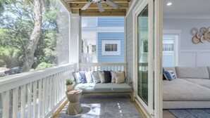 A balcony with a wooden ceiling fan, a cushioned seating area with a variety of pillows, and a view of a blue house through a glass door. - 69 Heated Pool | Kids Retreat | Near Beach | Sleeps Big (Hilton Head Island)