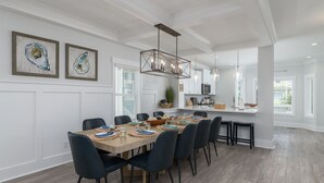 A modern dining room with a wooden table set for six, surrounded by dark chairs. There are two framed artworks on the wall, a kitchen area with white cabinets, and a hanging light fixture above the table. - 69 Heated Pool | Kids Retreat | Near Beach | Sleeps Big (Hilton Head Island)