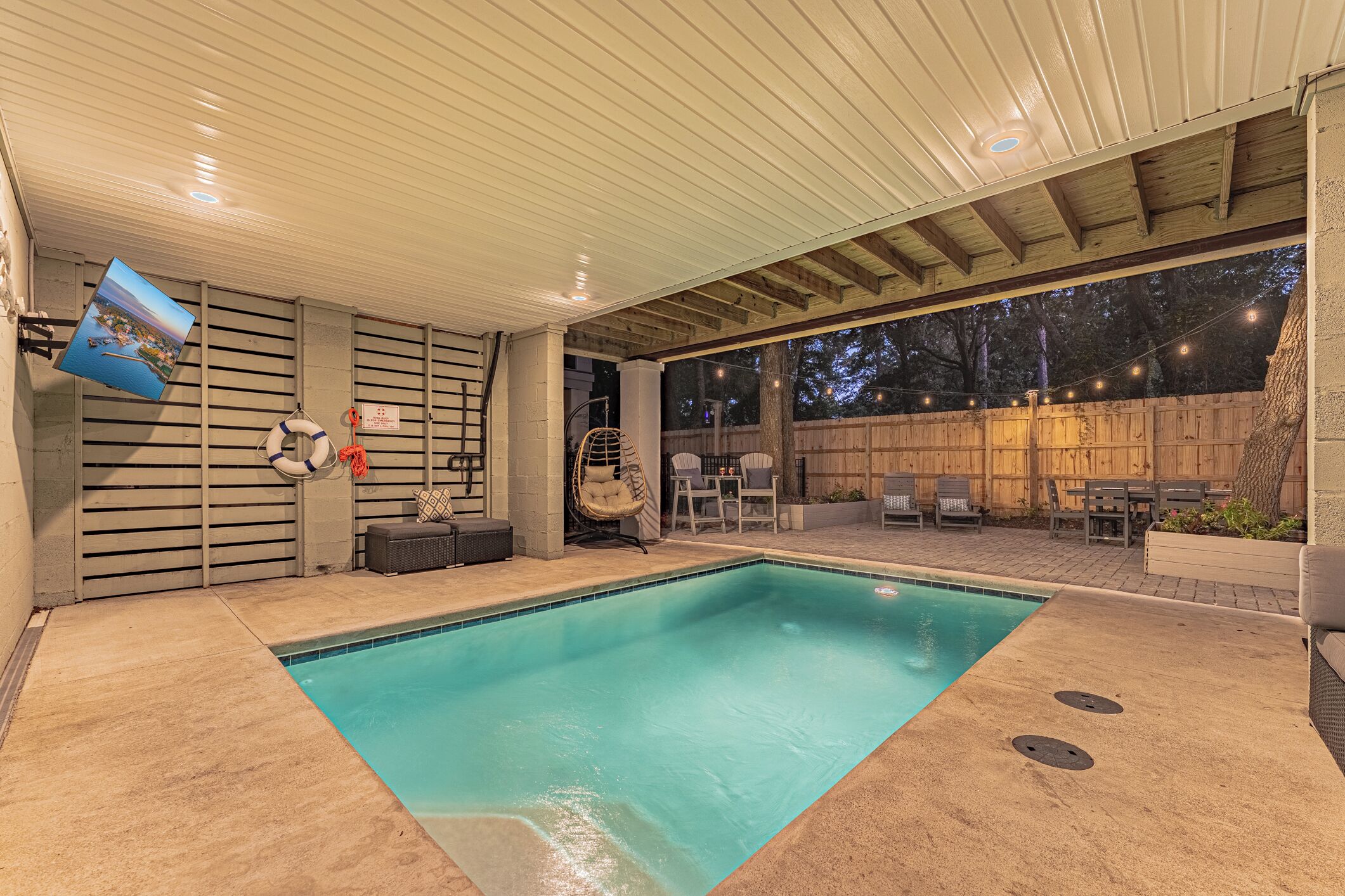 A swimming pool with a covered patio area, a hanging chair, and a view of the backyard.