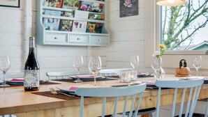 A dining area with a wooden table set for four, surrounded by blue chairs. There are wine glasses, a bottle of wine, and a vase with flowers on the table. Above the table, there are two pendant lights and a bookshelf filled with books and decorative items. - "Ylvie" - 150m from the sea by Interhome (Frørup)