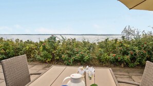 Une table dressée avec des croissants, des tasses à café et une théière, donnant sur un plan d’eau avec vue sur la mer et une rangée de plantes en fleurs.