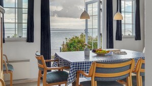 Un coin repas avec une table dressée pour deux personnes, deux lampes suspendues et une vue sur la mer grâce à de grandes fenêtres ornées de rideaux foncés.