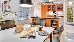 A modern kitchen with wooden cabinets, a dining table with chairs, a hanging light fixture, and a window with a view of greenery.