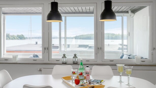 A modern dining area with a white table and chairs, two pendant lights, and a view of a body of water through large windows. - "Constance" - 50m from the sea by Interhome (Svendborg)