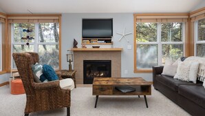 A living room with a fireplace, a television, and large windows. - Beachcomber Cottage - Short Distance to Beach 2 Bedroom - 2 Bath (Pacific City)