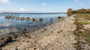 Una spiaggia con ciottoli e un molo di legno che si protende in acque calme, sotto un cielo sereno.