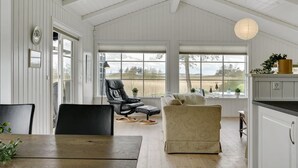 Une pièce spacieuse avec un plafond haut, une grande fenêtre donnant sur l’extérieur, une table à manger avec des chaises, un canapé et un coin cuisine avec des placards.