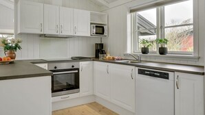 Une cuisine moderne avec des meubles blancs, un four encastré, un four à micro-ondes et un lave-vaisselle. Deux plantes en pot sont posées sur le rebord de la fenêtre et un panier de fruits se trouve sur le plan de travail.