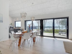 Et moderne spiseområde med et stort bord, hvide stole og udsigt til en sø gennem store skydedøre.