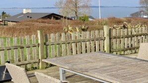 Eine Holzterrasse mit Tisch und Stühlen, mit Blick auf ein Gewässer und Gebäude im Hintergrund.
