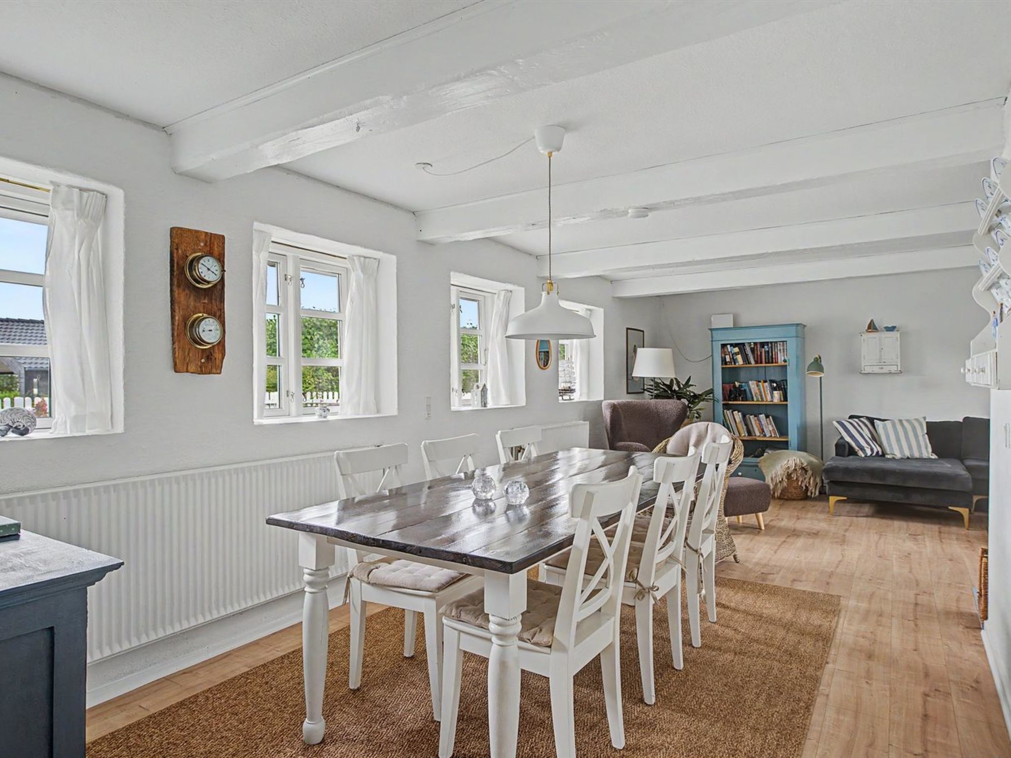 A dining area with a wooden table and white chairs, a blue sofa, and a bookshelf in a room with white walls and a wooden floor.