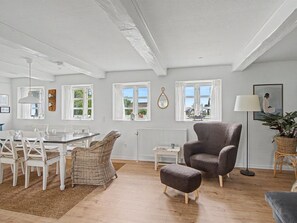 A room with a dining table, chairs, a grey armchair, a small ottoman, a floor lamp, and a potted plant.