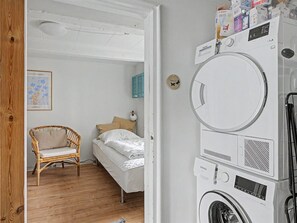 Une buanderie avec des machines à laver et des sèche-linge superposés, une chaise en osier, un lit avec du linge de lit blanc et un luminaire fixé au mur.