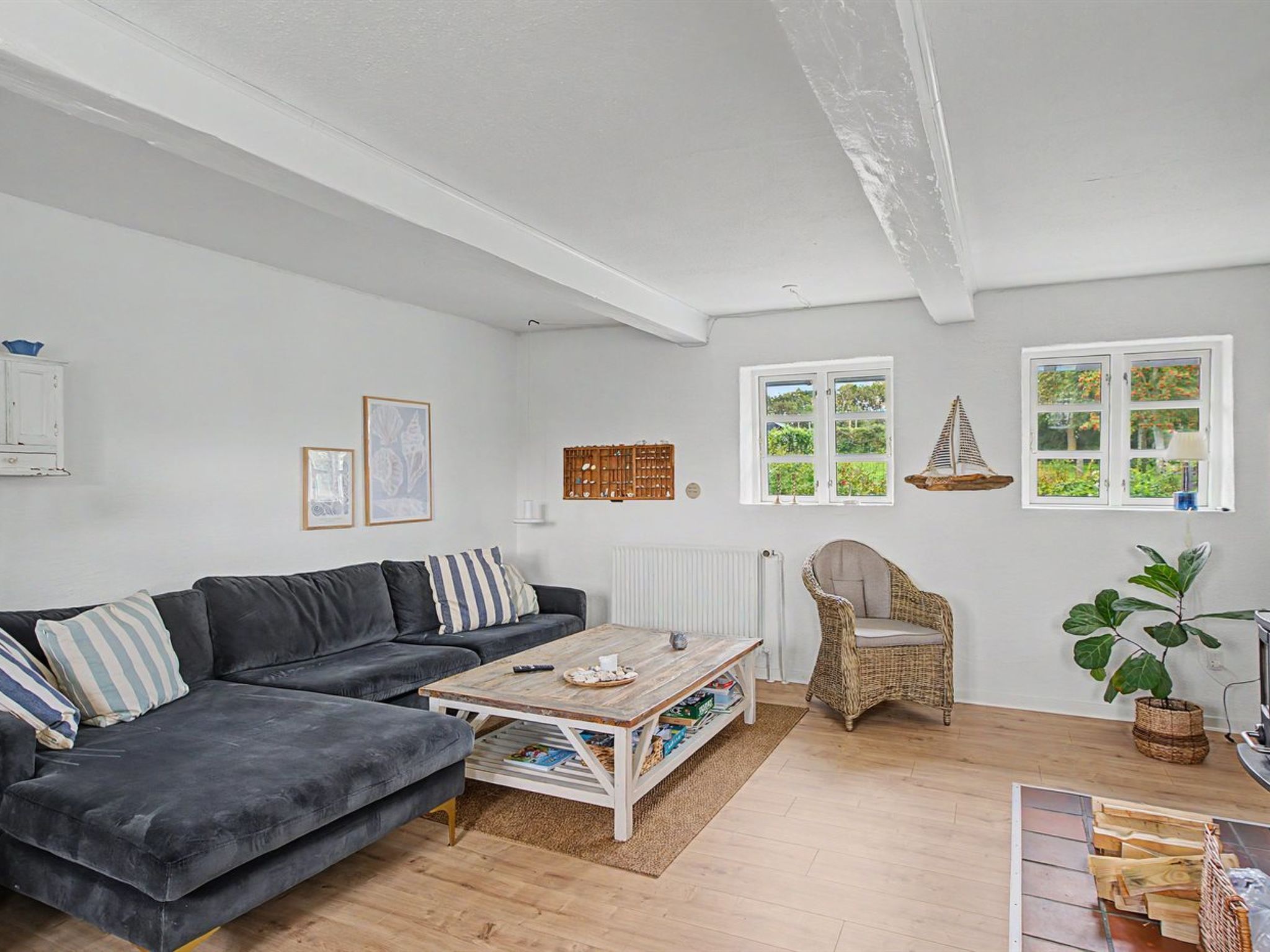 Un salon avec un canapé gris foncé, une table basse en bois, un fauteuil en osier et une grande fenêtre donnant sur un paysage verdoyant.