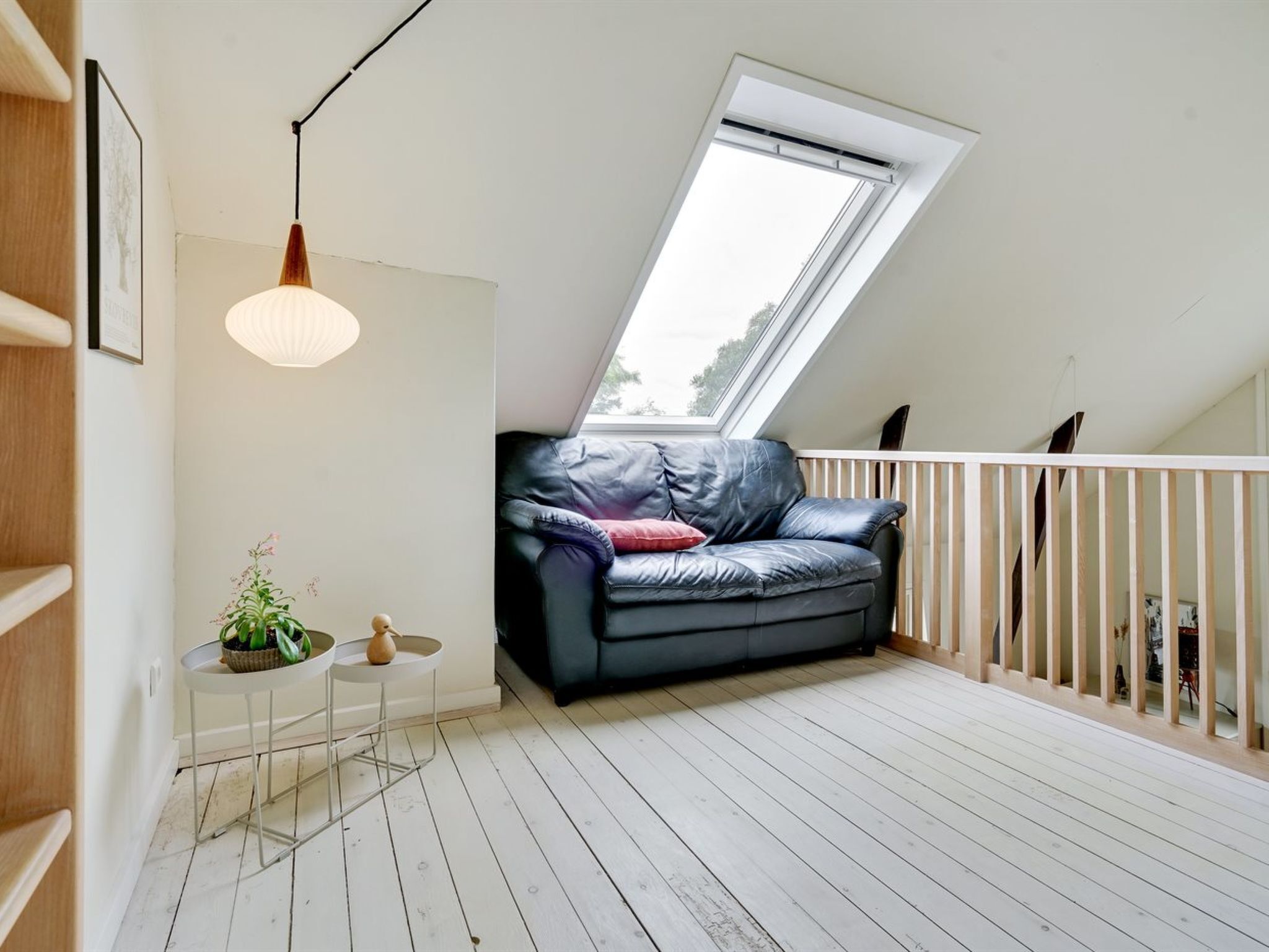 Ett sovrum på entresolplan med ett sluttande tak, en svart lädersoffa, ett litet vitt bord med en växt och en hängande lampa.