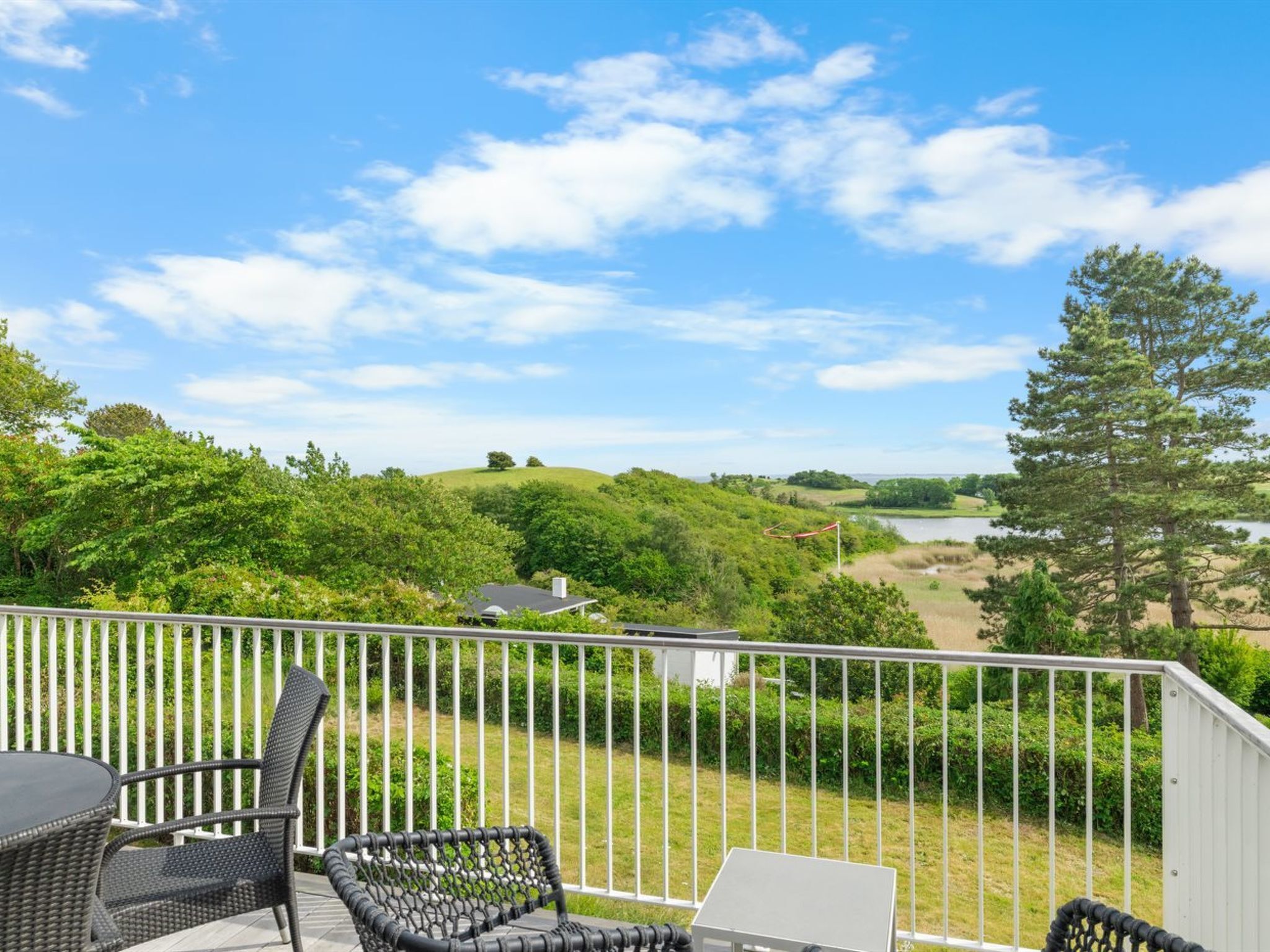 Un balcon doté de meubles en osier et d’un ordinateur portable, offrant une vue sur un paysage verdoyant avec des collines et un lac.