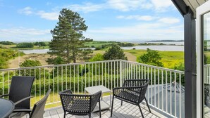 Un balcon avec des fauteuils et une table en osier, donnant sur un lac et des champs verdoyants.