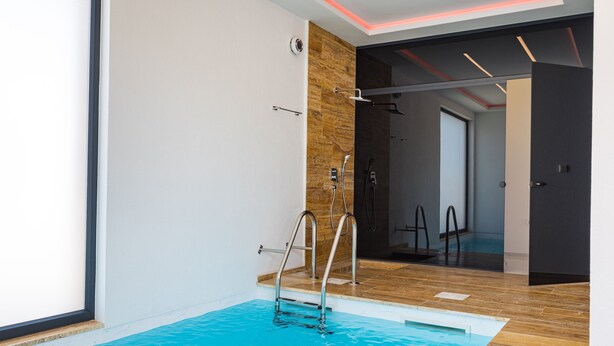 Piscina interior moderna com piso de madeira, parede de vidro e escada de metal.
