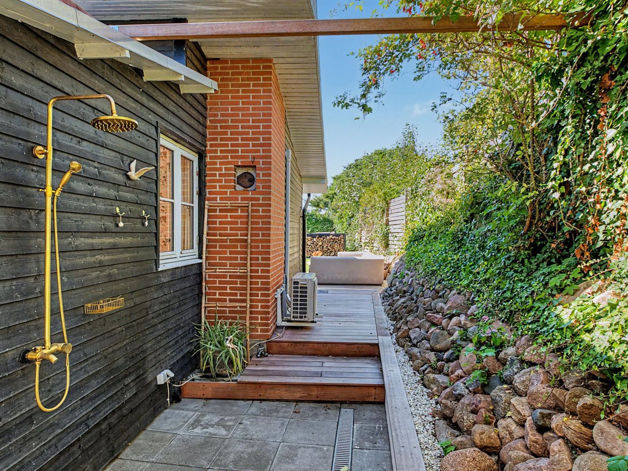 A residential property with a wooden exterior, a brick chimney, a metal water spigot, a stone pathway, and a garden with a stone wall and greenery.